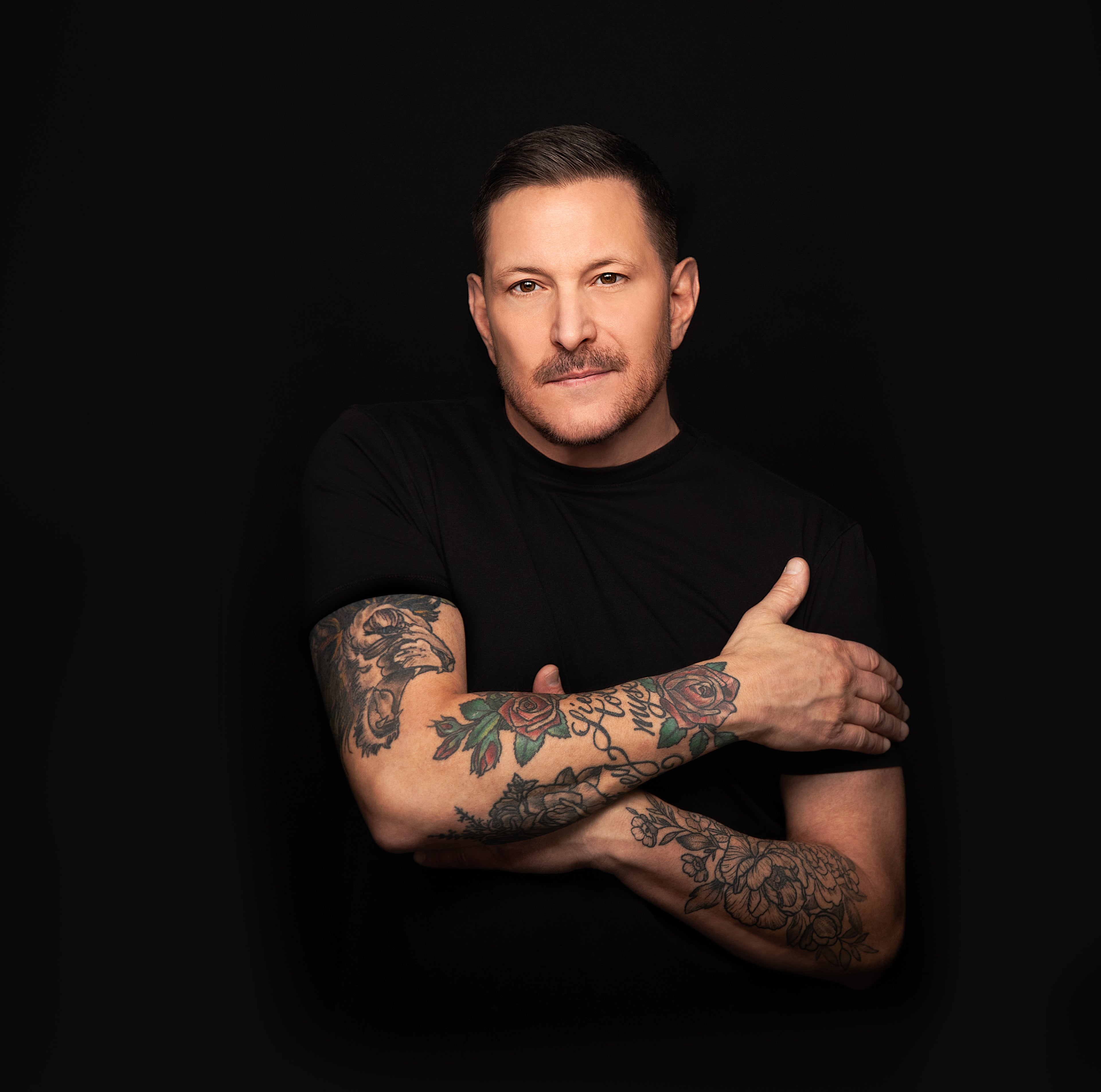 Man with tattoos on his arms wearing a black shirt against a dark background